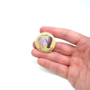 Pinback button featuring a design that inspires racial equality. Two hands of different skin color with a heart in the center. the text reads "Our magic is stronger together"
